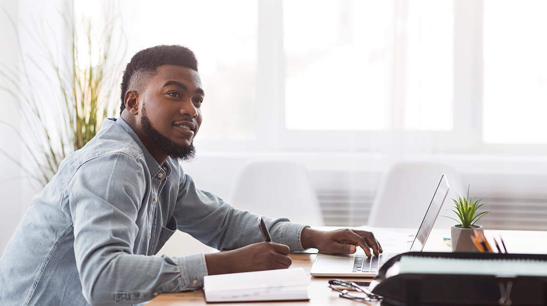 Young black man sitting at desk with laptop in a brightly lit, modern office. He has a trimmed beard, wears a pale denim shirt and is looking up and smiling