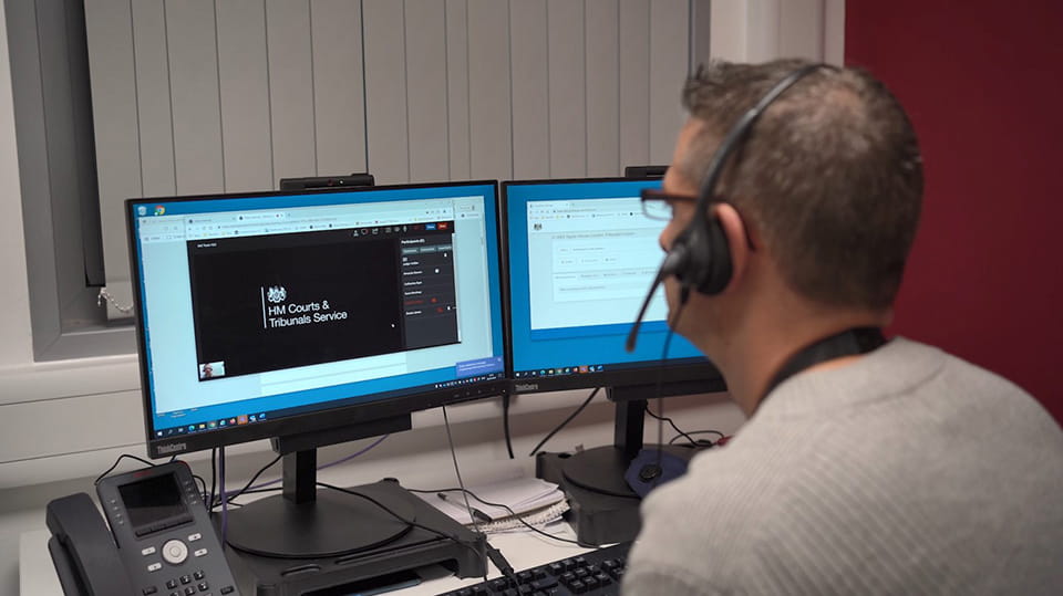 A court user waits for an online hearing to start; a white man wearing a headset looks at a computer monitor on his desk, the screen shows a video call holding screen with the HM Courts and Tribunals Service logo in white on a black background