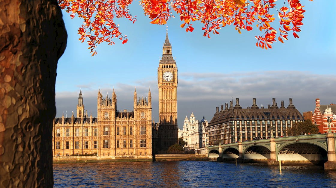 View of Westminster Bridge, Houses of Parliament and Big Ben across the Thames on a bright autumn day, with orange leaves and tree in the foreground