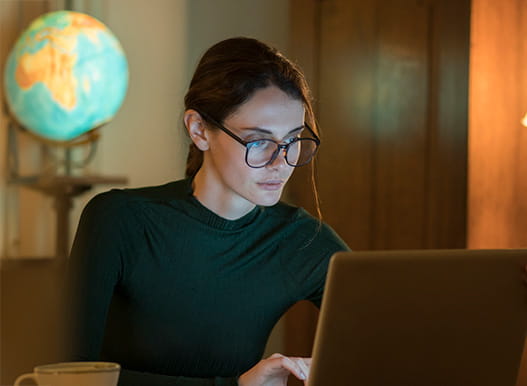 A woman wearing glasses and her hair tied back, looks at her laptop in a dark room with a globe in the background.