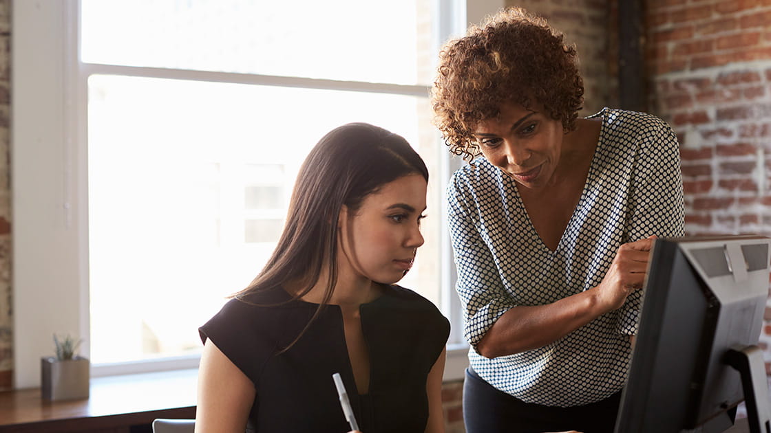 Solicitor advises young colleague on her work, standing by her desk while they look at computer screen together